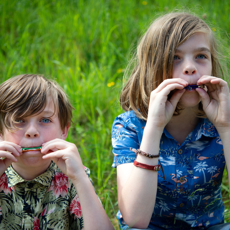 Two young boys sitting out on the grass blowing into harmonicas they have made