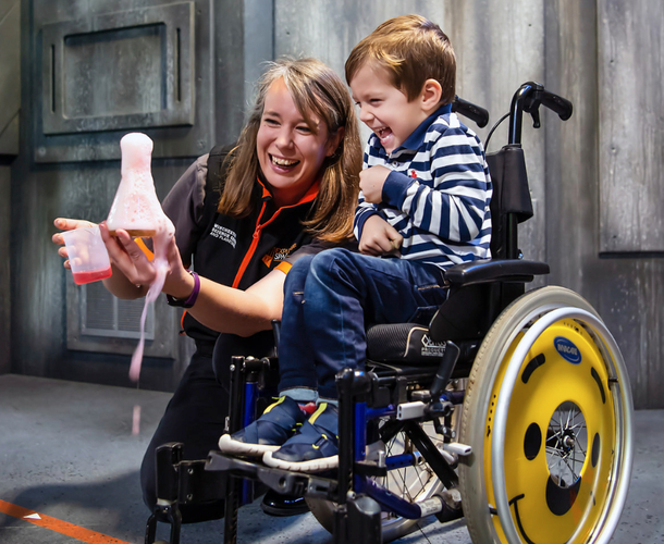 A young boy laughing in a wheelchair with a smiling presenter crouched beside him, holding a beaker containing a chemical reaction