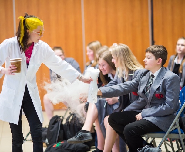 A group of school children looking at a beaker which has smoke coming out of it being held by a presenter in a lab coat