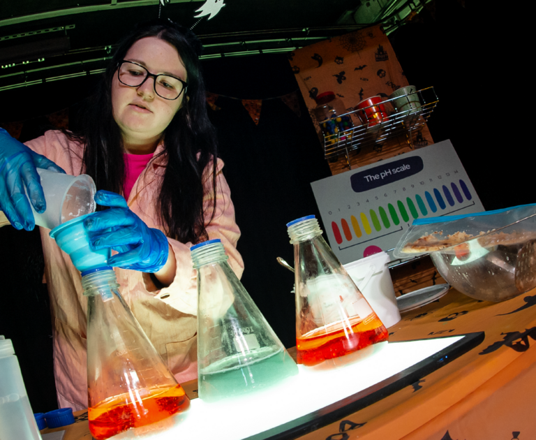 Aly, Public Curiosity Officer at Winchester Science Centre is wearing a white lab coat and blue rubber glovers. She is pouring chemicals into beakers.