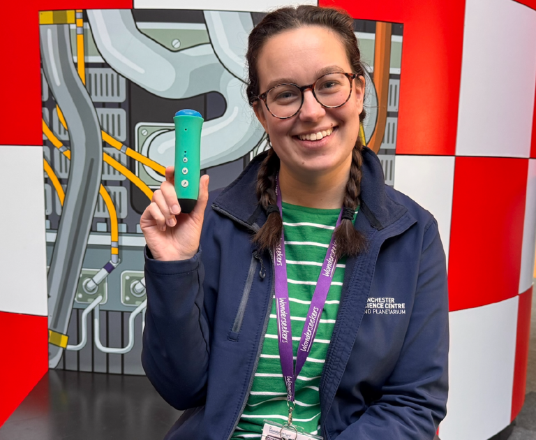 A women is sat on the large rocket exhibit, smiling. She has long dark brown hair that's been platted and is wearing her navy-blue Winchester Science Centre jacket. In her left hand is a Discovery Pen.