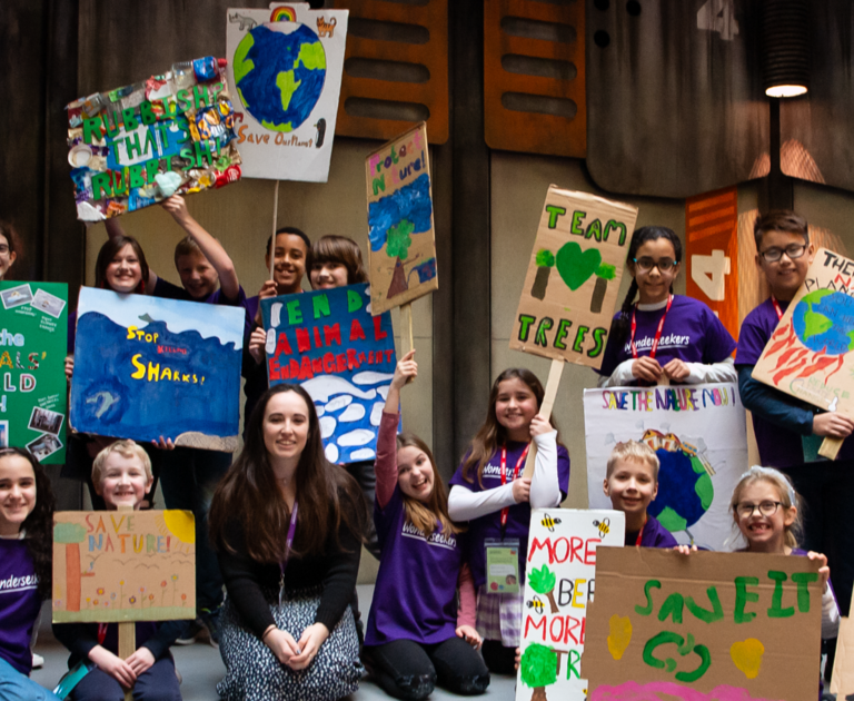 A group of children are holding homemade cardboard signs decorated with slogans about looking after wildlife and the environment.