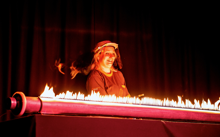 Live science show photo showing a female presenter smiling behind a tube lit up with flames