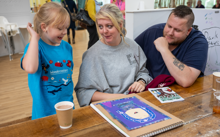 A young girls is showing her art book to her parents. They are all smiling.