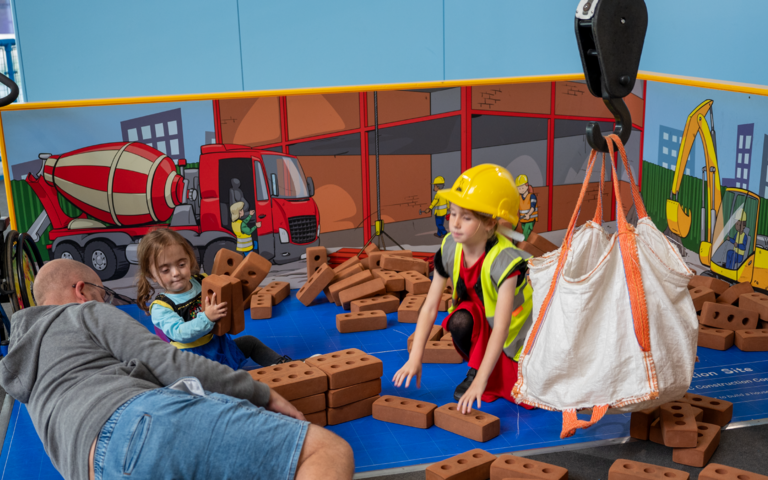 Two children are playing with foam bricks. One of them is playing with their parent and the other is wearing a yellow hardhat and a high visibility jacket.