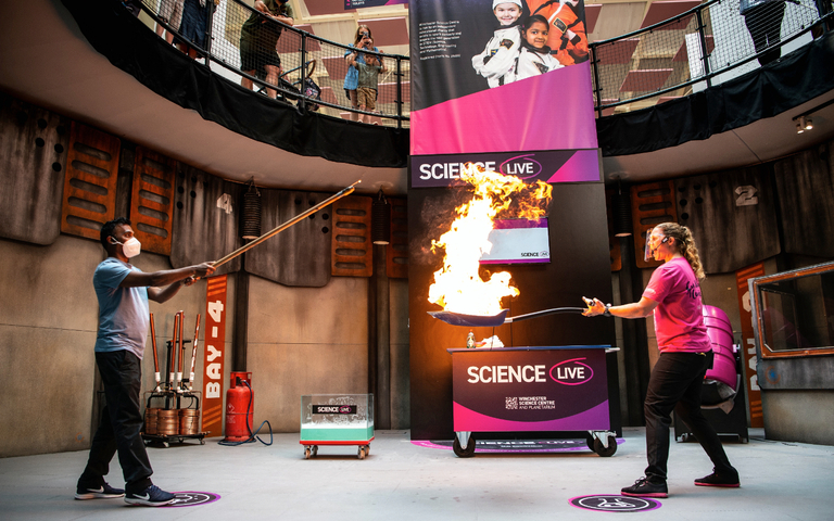 Live science demo area photo showing a presenter holding a spade with liquid that's on fire, and a visitor holding a large stick used to light the fire
