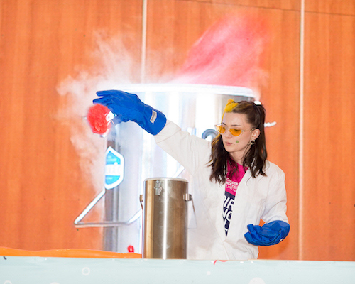 A presenter in a lab coat, gloves and safety goggles holding something that is filling the room with smoke