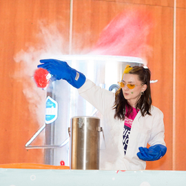 A presenter in a lab coat, gloves and safety goggles holding something that is filling the room with smoke