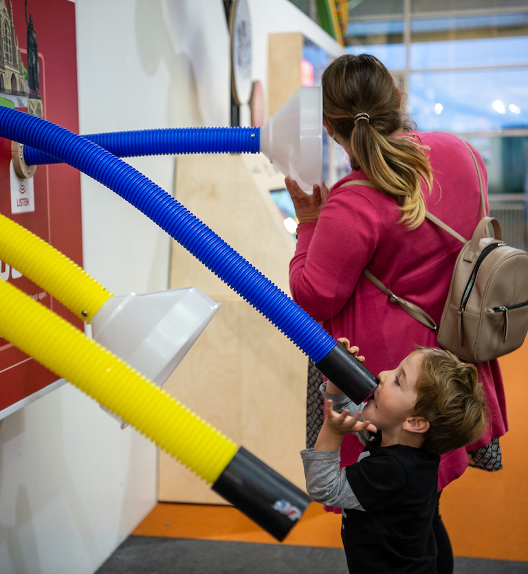 Interactive science exhibition photo showing a young boy talking into a tube while his mum holds her ear to the other end to listen
