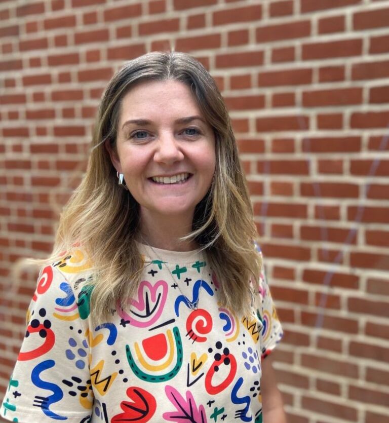 A woman with long blonde hair, wearing a cream t-shirt with colourful, squiggly patterns on it, is standing in front of a red brick wall and smiling directly at the camera.
