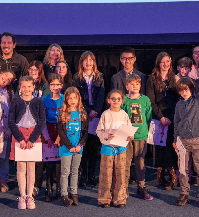 A group pf people are stood on a stage in front of a Planetarium screen. Adults are at the back and children are at the front. Some of the children are holding certificates.
