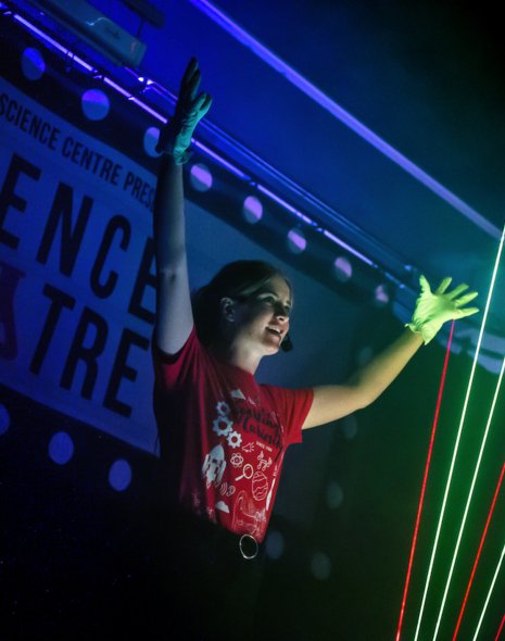 Live science show photo showing a female presenter with her hands in the air showing off the beams of light from a laser harp